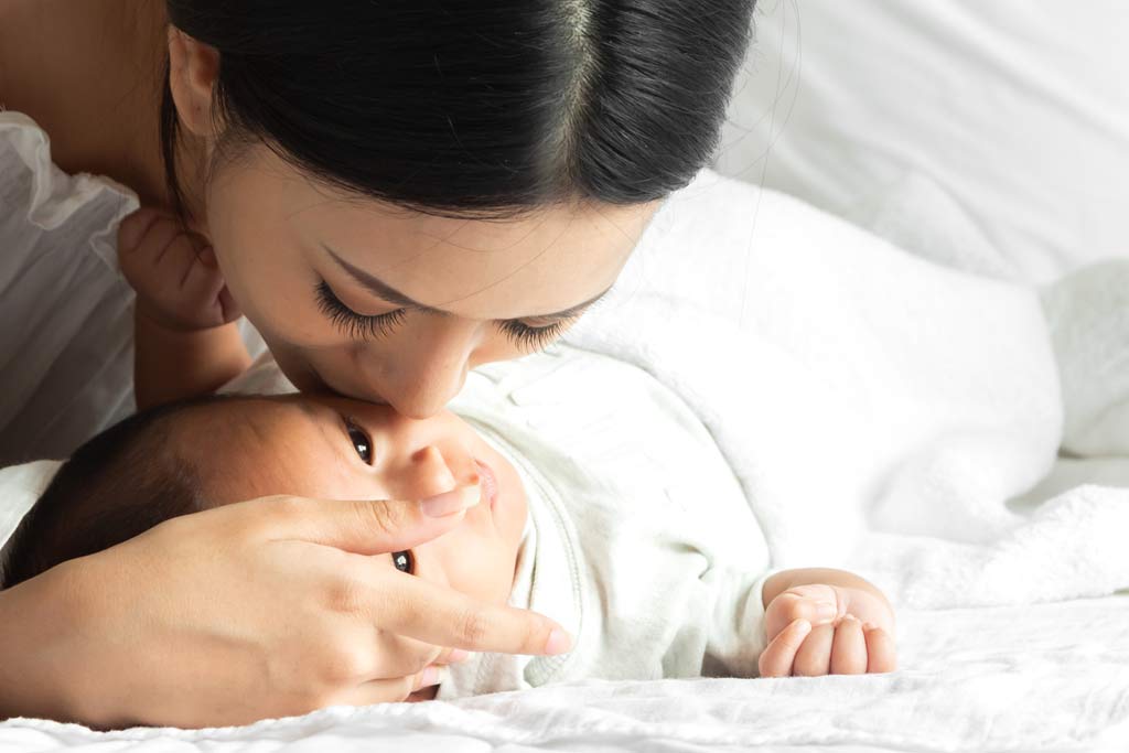 mom kissing baby lying down with white sheets in background